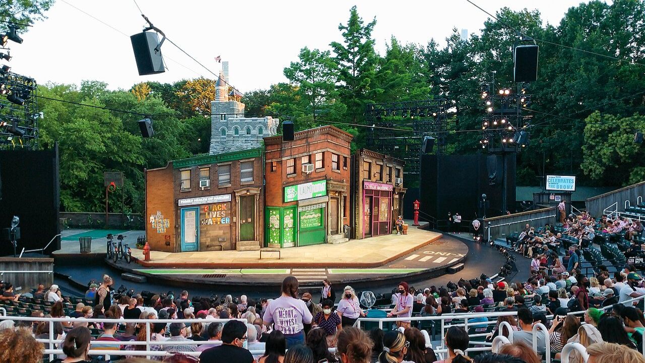 Audience watching outdoor Shakespeare in the Park performance with urban-themed stage set.