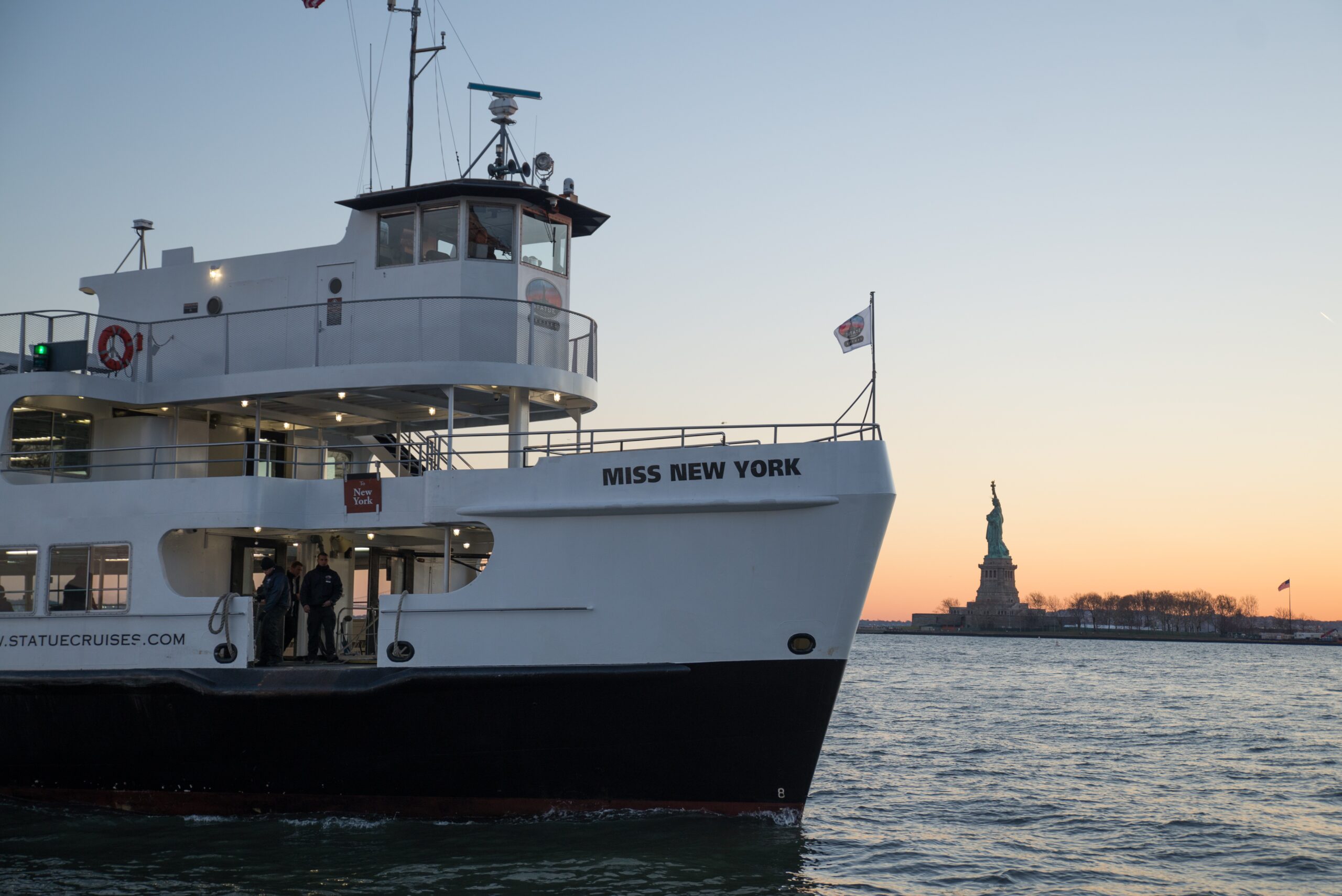 Cruise boat moving across New York Harbor with the Statue of Liberty visible far in the distance and open water surrounding the scene under soft evening light.