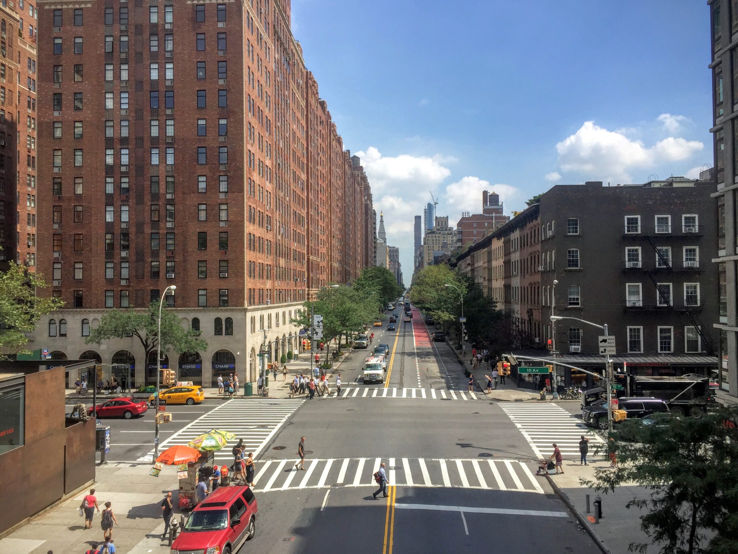 Aerial view of busy Manhattan streets during summer, with yellow taxis, tall skyscrapers, green trees, and sunlight reflecting across the city roads.