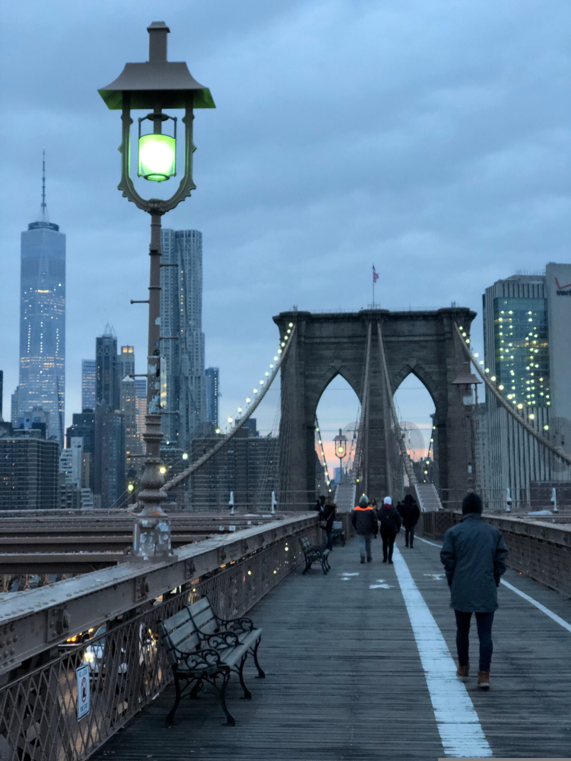 People walking across the Brooklyn Bridge at dusk with glowing skyline views, soft evening lights, and the Manhattan cityscape in the background.