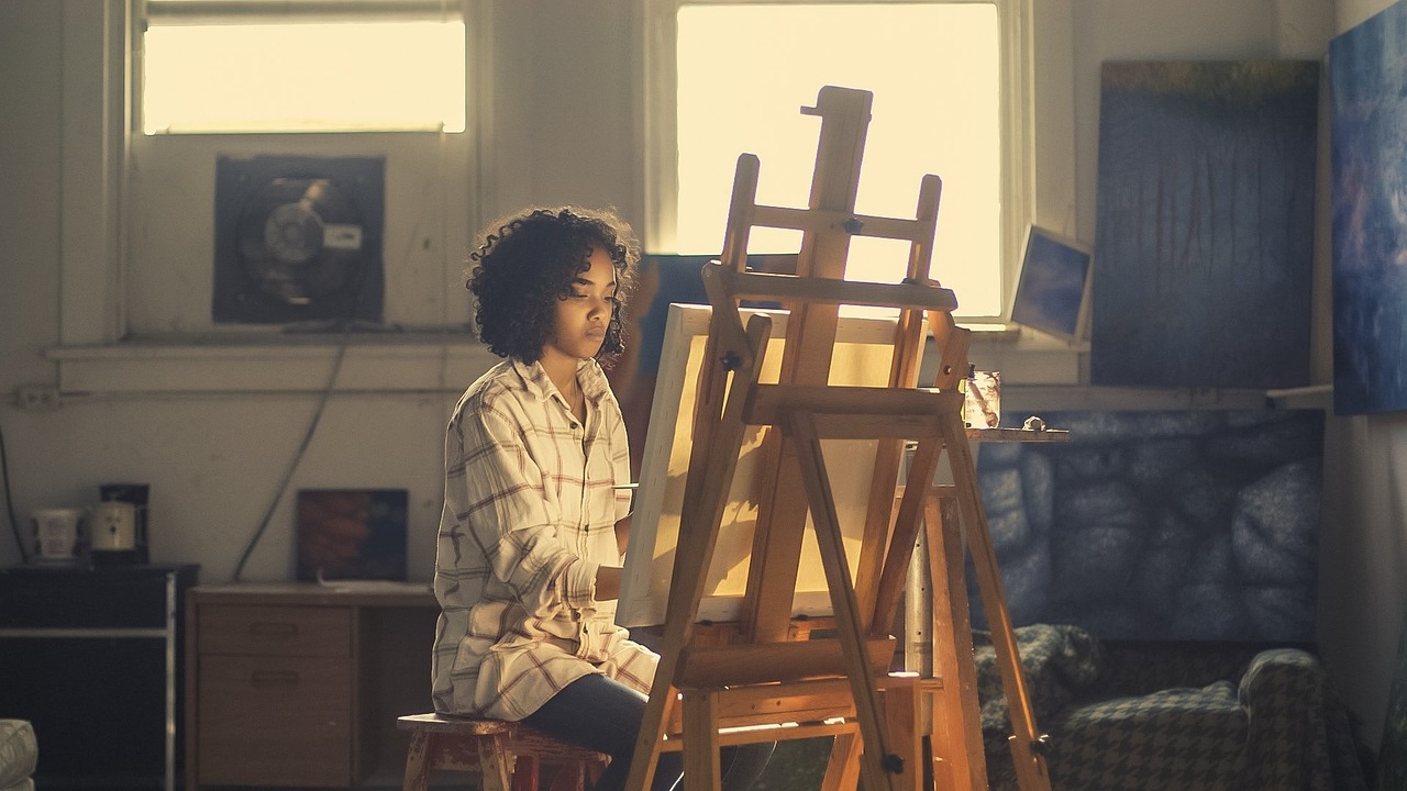 Young artist painting at an easel near sunlit windows.